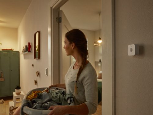 Person carrying laundry basket in a hallway, with a white smart sensor on the wall and warm pendant lighting in the background.