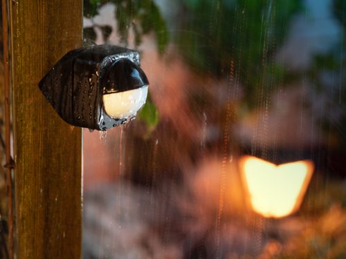Outdoor smart sensor mounted on a wooden post, covered with raindrops, designed to detect motion for smart security lighting control.