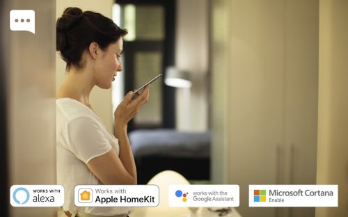 Person holding a smartphone and speaking into it, with logos for Alexa, Apple HomeKit, Google Assistant, and Microsoft Cortana shown.