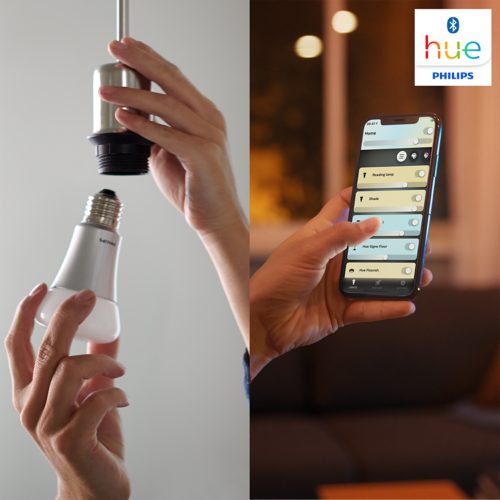 Person installing a white smart light bulb and using a smartphone app to adjust lighting settings hue logo visible on screen.