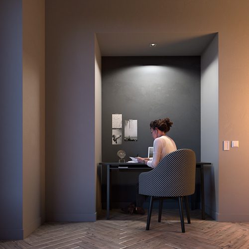 Person seated at a desk in a home office alcove, writing under focused ceiling lighting, with wall art and a small lamp visible.