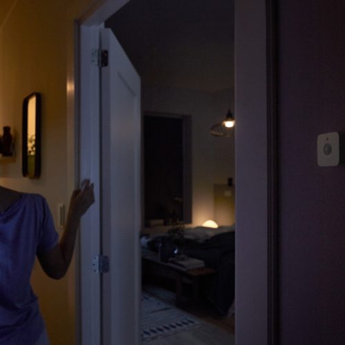 Person entering a dimly lit bedroom, with a wall-mounted sensor visible and warm lighting illuminating the room in the background.