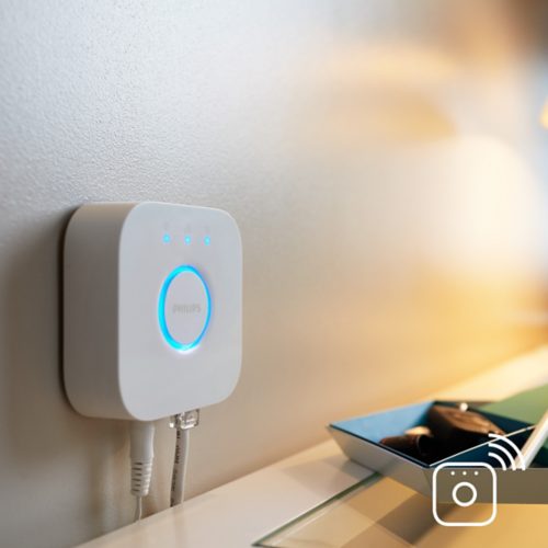 White smart hub mounted on wall above desk, illuminated blue ring and status lights, network cables connected, tray nearby.