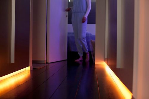 Person walking barefoot in a hallway at night, illuminated by warm floor-level lighting along the base of the walls.