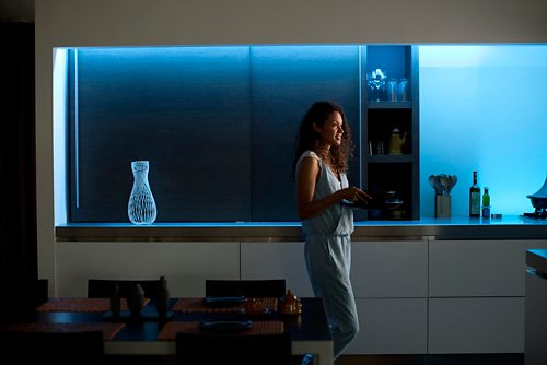 Person standing in a modern kitchen holding dishes, with blue smart lighting illuminating the countertop and shelves.