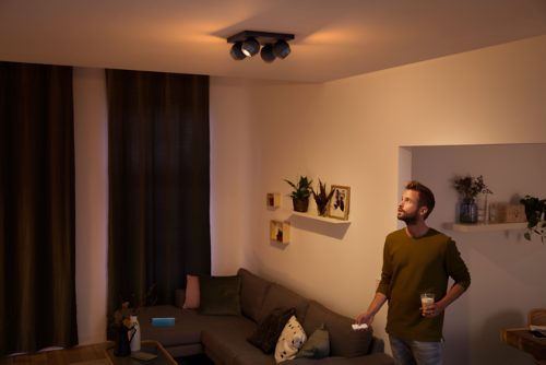 Person standing in a living room holding a drink and remote, with ceiling spotlights illuminating the seating area.