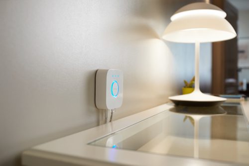 Square white smart hub mounted on wall above a glass desk, with illuminated blue ring and cables connected at the bottom.