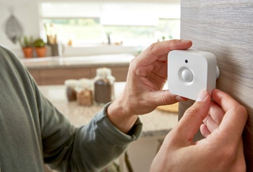 Hands mounting a white motion sensor onto a wooden wall, likely for smart home security or lighting control purposes.