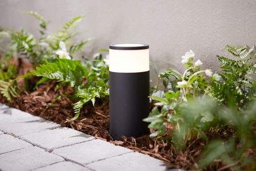 Black cylindrical outdoor light fixture installed in a garden bed, illuminating nearby plants and adjacent stone pathway.