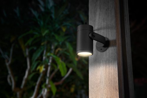 Black outdoor wall-mounted spotlight illuminating a wooden post, with surrounding plants visible in the background.