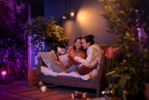 Two people sitting on a patio sofa sharing a drink, illuminated by adjustable wall-mounted smart lights in a cozy outdoor setting.