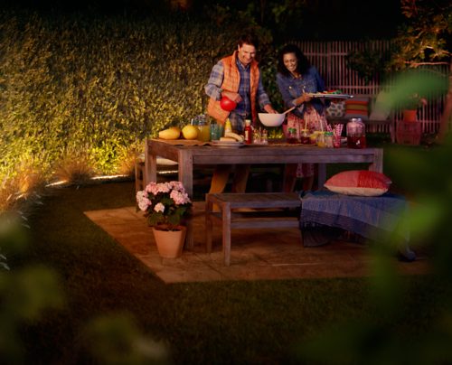 Two people preparing food at a garden table in the evening, with outdoor lighting illuminating the greenery and dining area.