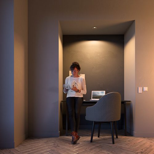 Person reading a book while standing in a home office nook, illuminated by overhead lighting above a desk with a laptop.