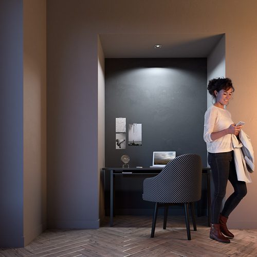 Person standing near a home office desk with focused ceiling light illuminating workspace, laptop, and wall decor in a modern setting.