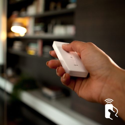 Hand pressing button on a white smart lighting remote control, likely adjusting brightness or turning lights off in a living room.