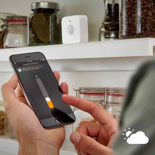 Finger adjusting daylight sensitivity slider on a smartphone app, with a motion sensor device visible on a white shelf in the background.