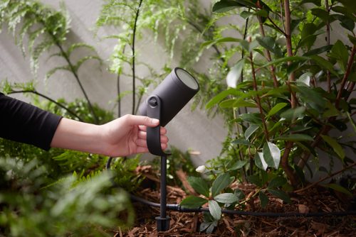Hand adjusting a black outdoor spotlight in a garden bed among plants, focusing the light fixture for landscape illumination.