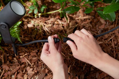 Hands connecting two black outdoor cable connectors to a spotlight fixture, positioned on garden mulch near green plants.