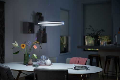 Circular pendant light in metallic finish suspended above dining table, illuminating tabletop with flowers, glassware, and magazines.