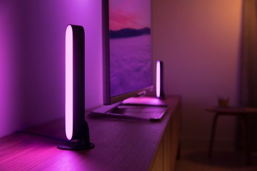 Vertical smart light bar placed on a cabinet beside a monitor, emitting purple light onto the wall and surface.