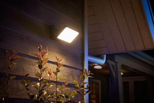 Square outdoor wall light fixture mounted on exterior siding, illuminating nearby plants and wall surface.