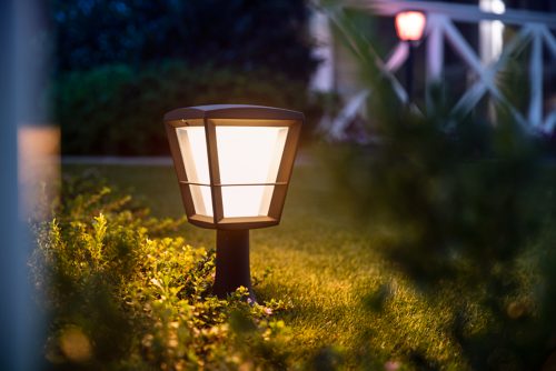 Outdoor lantern-style smart light fixture installed in a garden bed, casting white illumination onto surrounding grass and plants.