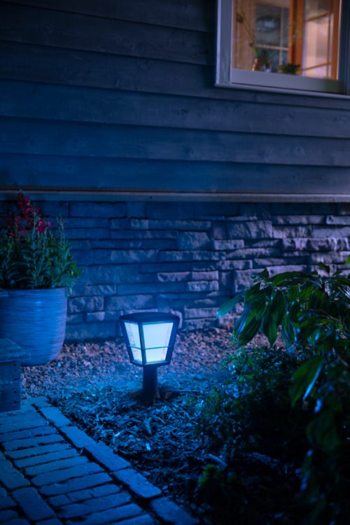 Outdoor smart lantern with a dark frame installed along a garden path, emitting blue light onto nearby plants and stone wall.