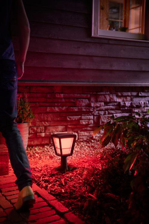 A person walking on a garden path at night, illuminated by a red outdoor smart lamp near a house with brick siding.