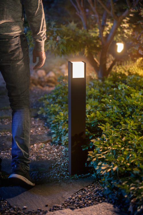 Person walking on a garden path illuminated by a modern outdoor bollard light, with greenery and trees visible in the background.
