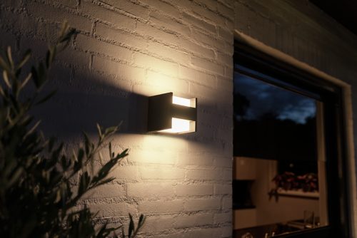 Wall-mounted rectangular outdoor light fixture emitting illumination onto a white brick exterior wall near a window and foliage.