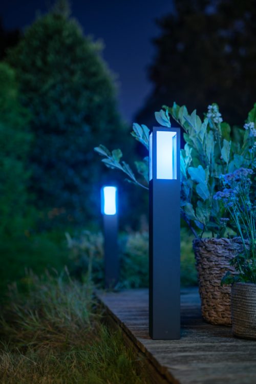 Outdoor black bollard lights installed along a wooden garden path, emitting blue light near potted plants and greenery.