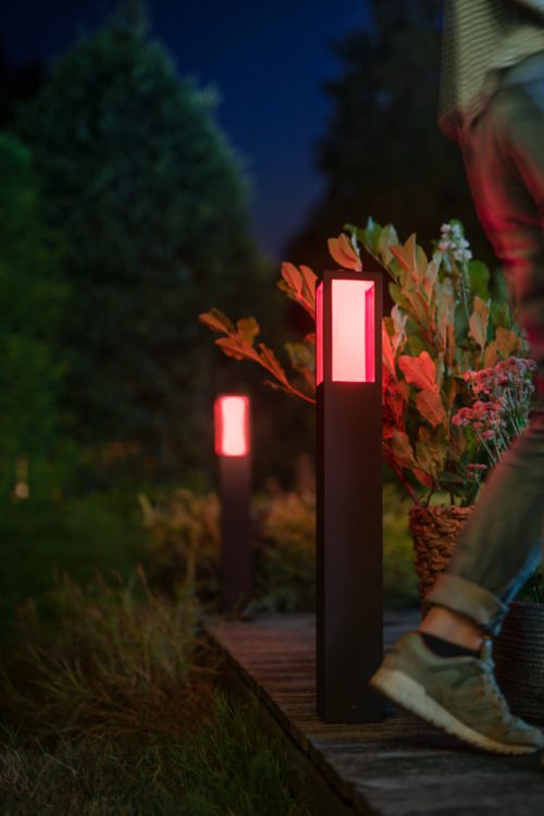 Person walking on a garden pathway at night illuminated by outdoor lights with a red glow, surrounded by plants and greenery.