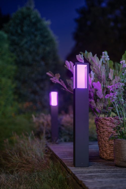 Black outdoor bollard light installed along a wooden garden path, emitting purple illumination near plants and baskets.