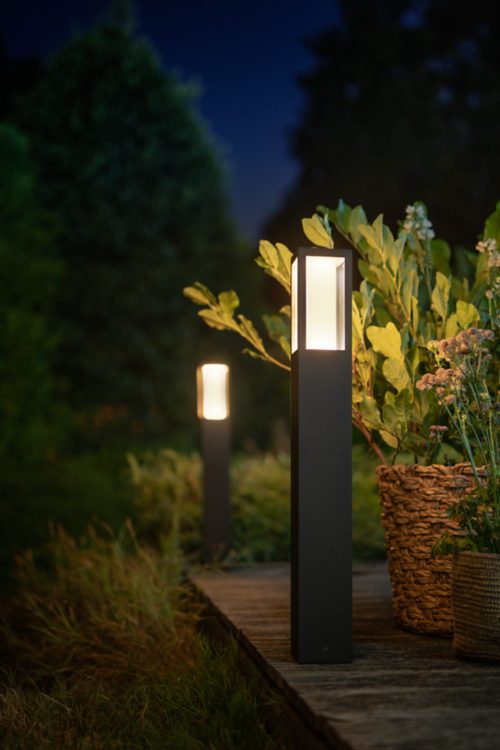 Outdoor smart lighting bollard with rectangular design installed along a wooden pathway, illuminating nearby plants at night.