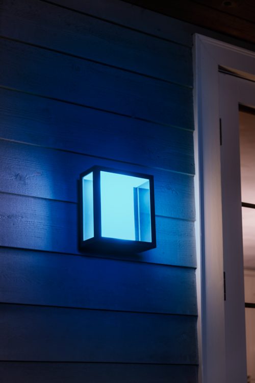 Square outdoor wall light emitting blue illumination, mounted beside a door on horizontal wood siding, casting colored light on surface.