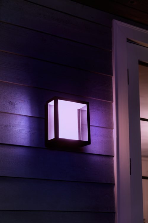 Square outdoor wall light fixture mounted beside a door, emitting purple illumination onto horizontal wood siding.