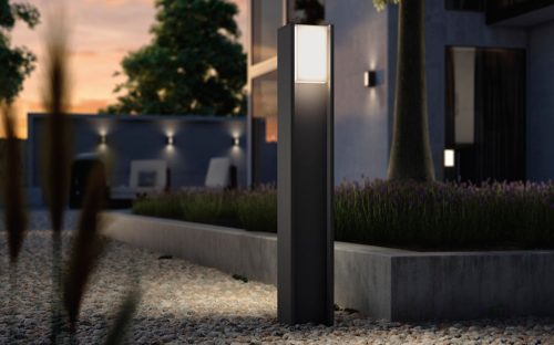 Outdoor smart bollard light with rectangular design installed on gravel pathway, illuminating the surrounding garden area.