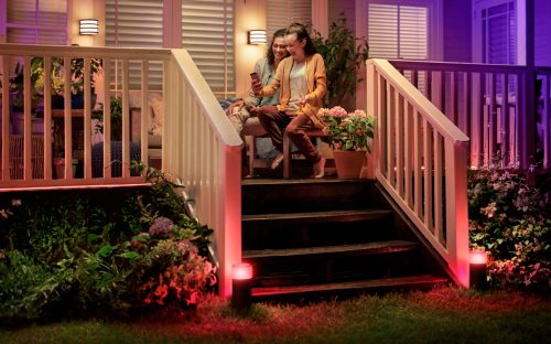 Two people sitting on a porch using a smartphone, with colored outdoor lights illuminating the garden and steps in the evening.
