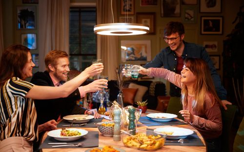 A group of people raise glasses in a toast over a dining table, illuminated by a circular pendant light in a home setting.
