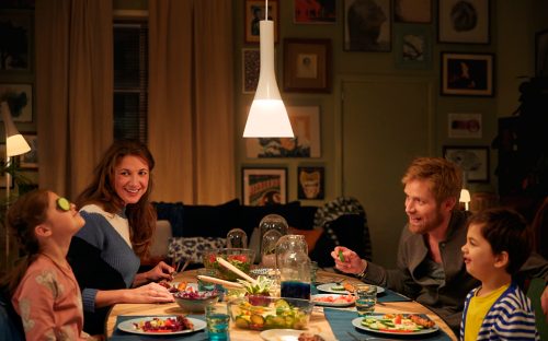 A group of people share a meal at a dining table under a white pendant light in a warmly lit living room setting.