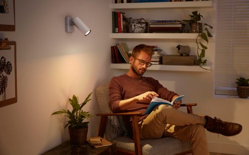 Person seated in a living room reading a book, illuminated by a wall-mounted spotlight with warm light.Shelves and plants visible.