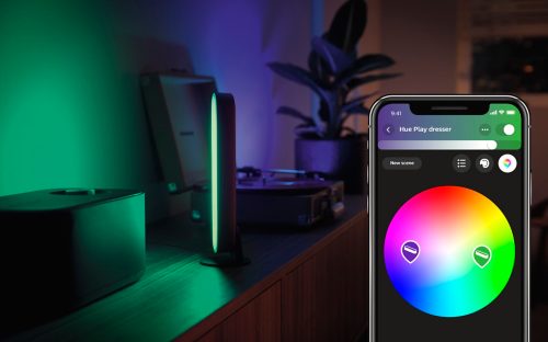 A smart light bar emits green light on a wooden surface, with a mobile app interface showing a color wheel and scene controls.