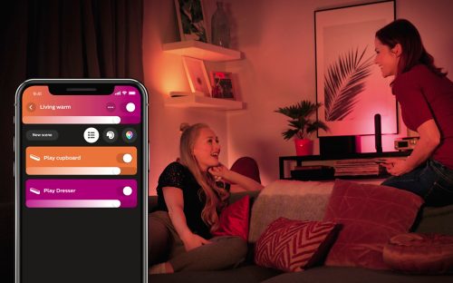 Two people in a living room adjust smart lighting with a mobile app, warm light illuminating shelves and decor in the background.
