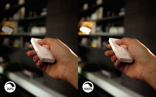 Hand pressing buttons on a white remote control to adjust lighting brightness, with 20% and 100% icons shown in the corners.