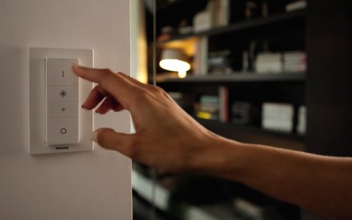 Finger pressing the top button on a white smart wireless light switch mounted on a wall, likely to turn on smart lighting.