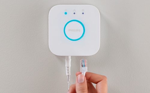 Hand holding an Ethernet cable near a white smart hub mounted on a wall, preparing to connect the cable to the device.
