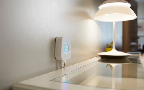 White smart hub mounted on wall above a glass-topped desk, illuminated with a blue ring, positioned near a table lamp.