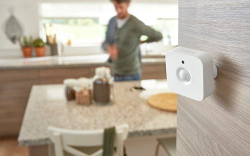 A white motion sensor device mounted on a wooden wall in a modern kitchen, with a countertop and jars in the background.