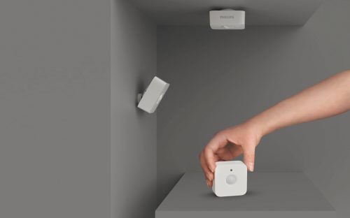 Hand placing a white motion sensor on a flat surface, with additional motion sensors mounted on the wall and ceiling.
