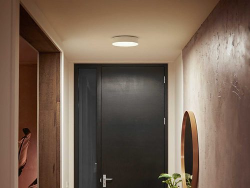 Ceiling-mounted round smart light fixture illuminating a hallway with textured walls, a dark door, and a mirror on the right wall.